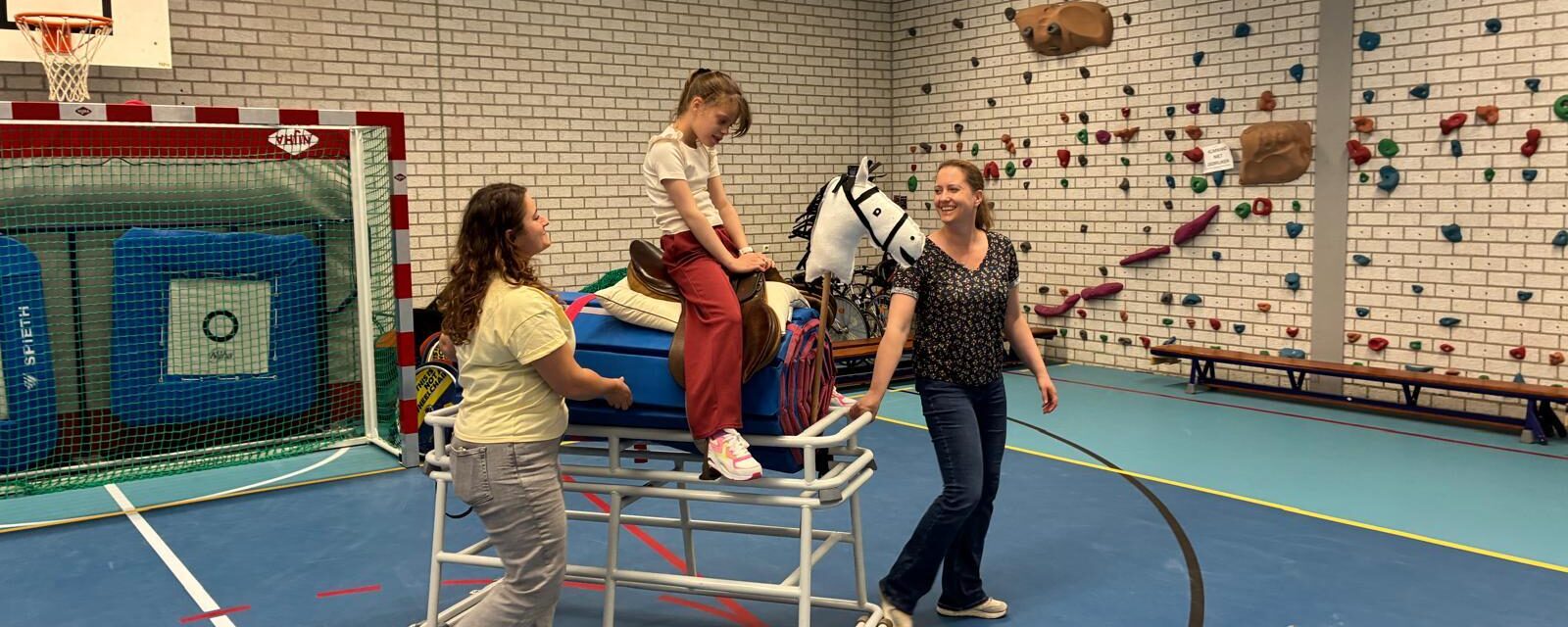 Tess is samen met twee begeleiders in de gymzaal en zit op een paard met wielen.