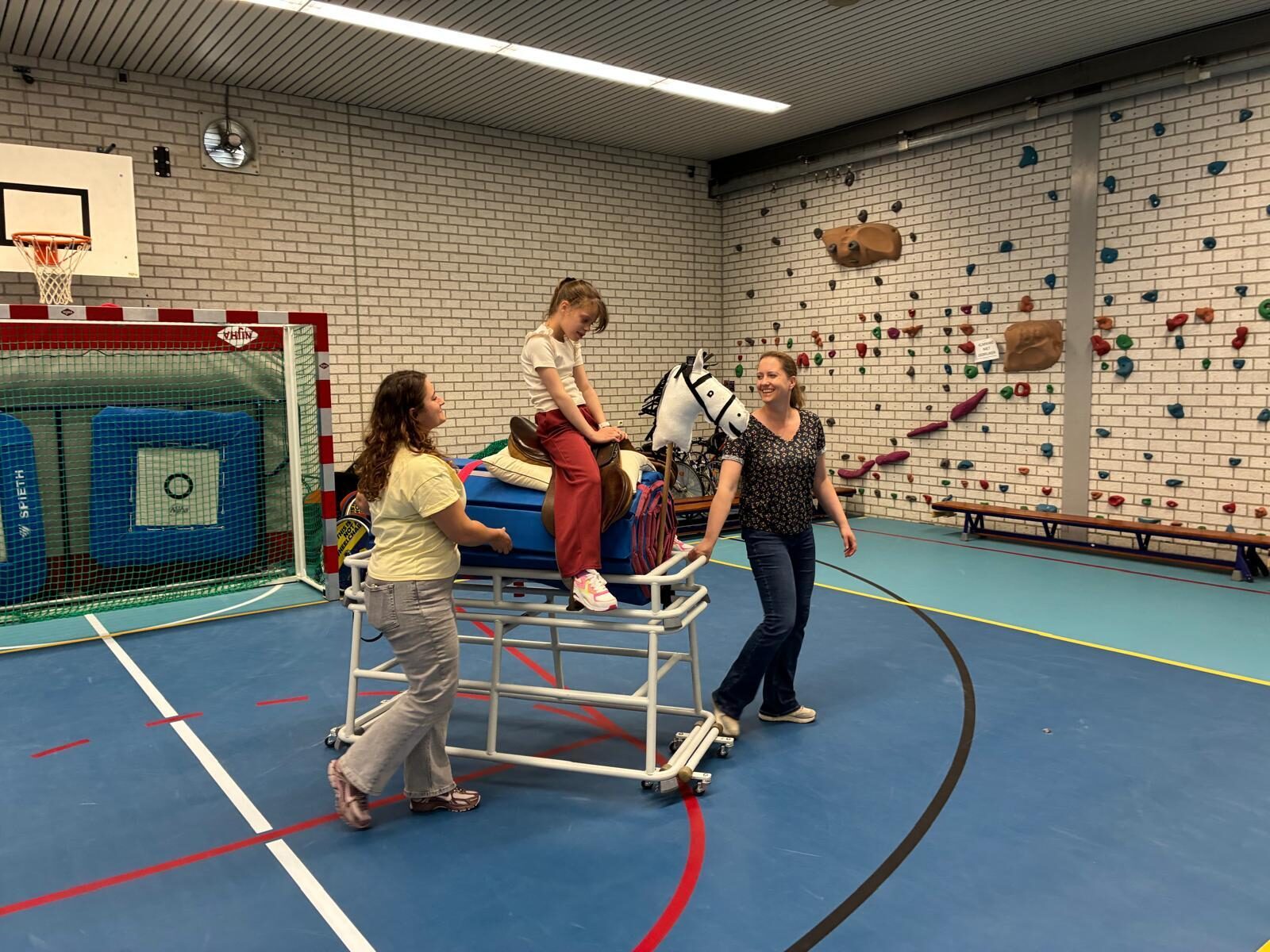 Tess is samen met twee begeleiders in de gymzaal en zit op een paard met wielen.