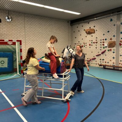 Tess is samen met twee begeleiders in de gymzaal en zit op een paard met wielen.